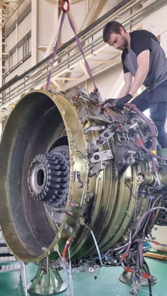 Technician inspecting a large aircraft engine in an industrial facility, with various mechanical components visible.